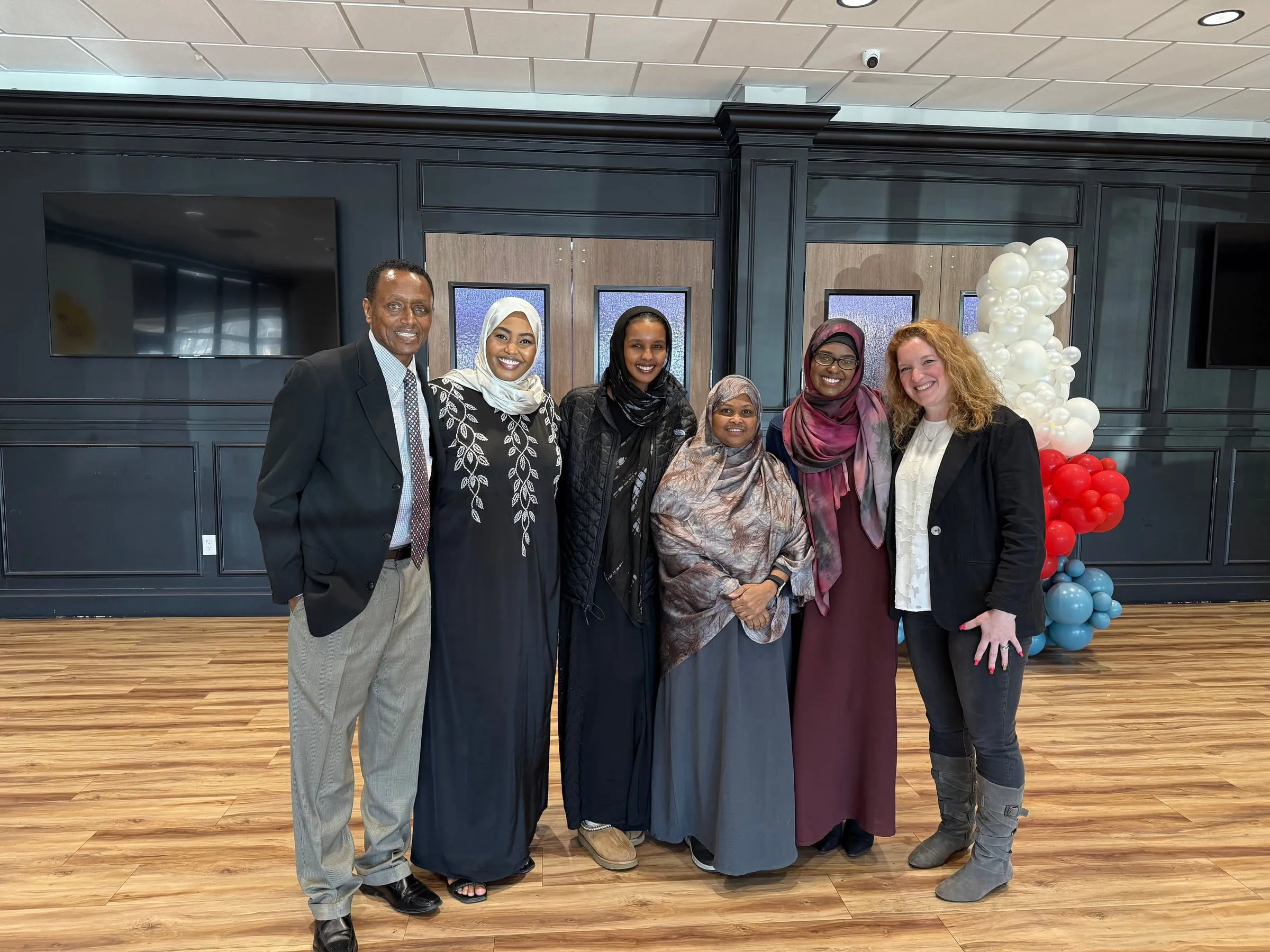 Maangaar Voices members stand with a man in a suit and a woman in a black blazer in an event hall decorated with balloons.