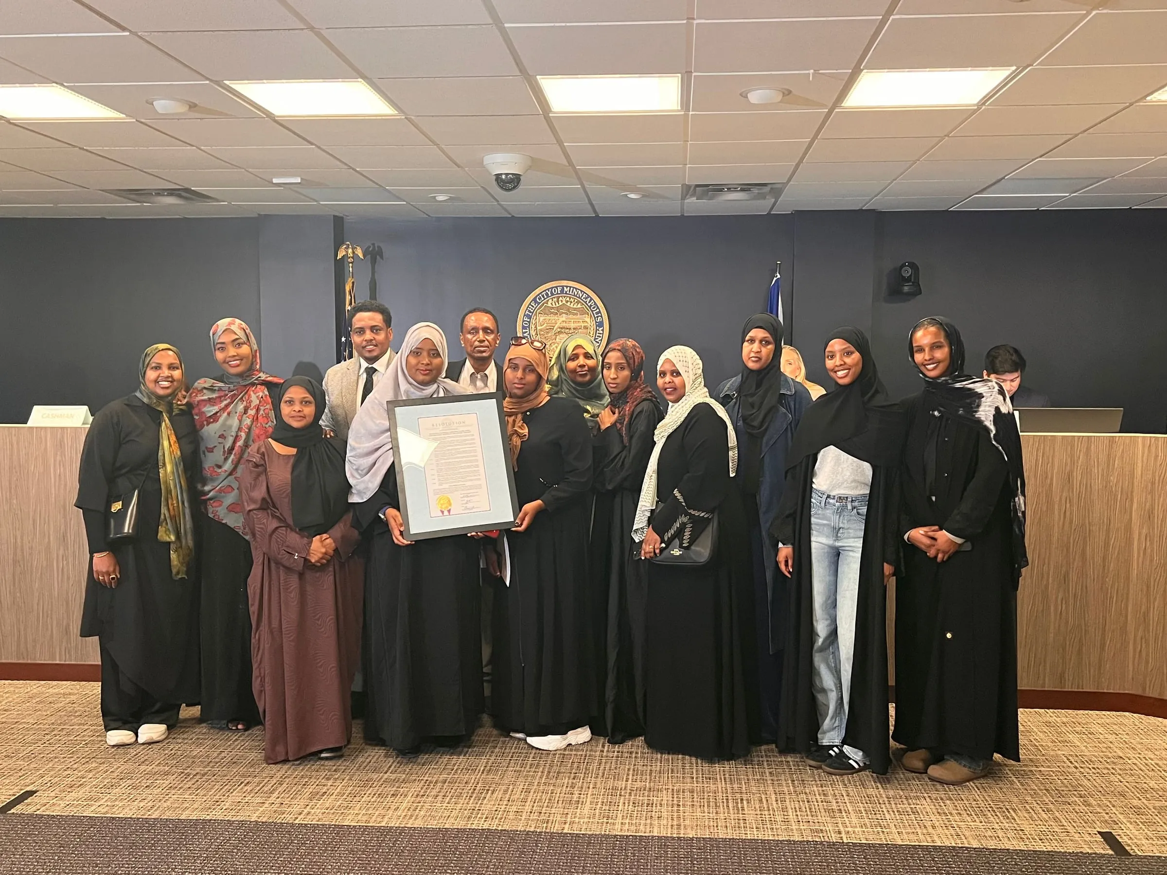 Maangaar Voices members and families hold the framed resolution in front of the City of Minneapolis seal in the City Council chambers.