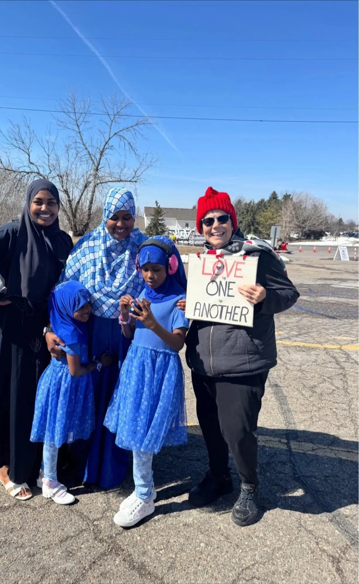 Two mothers and two girls in blue Eid dresses, one wearing pink ear defenders, beside a volunteer holding a “Love one another” sign outside the masjid.