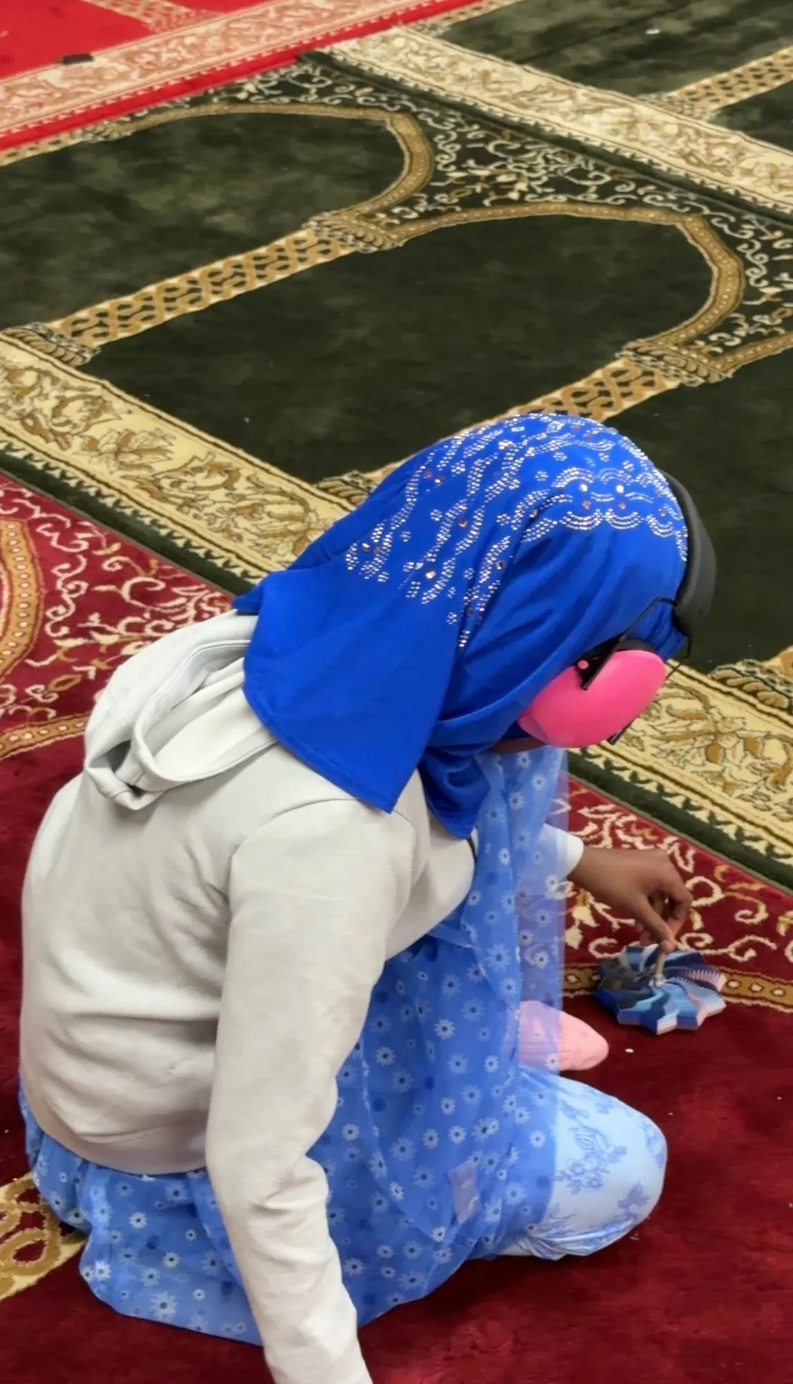 A girl in a blue hijab and pink ear defenders kneels on the masjid prayer carpet, playing with a fidget toy.