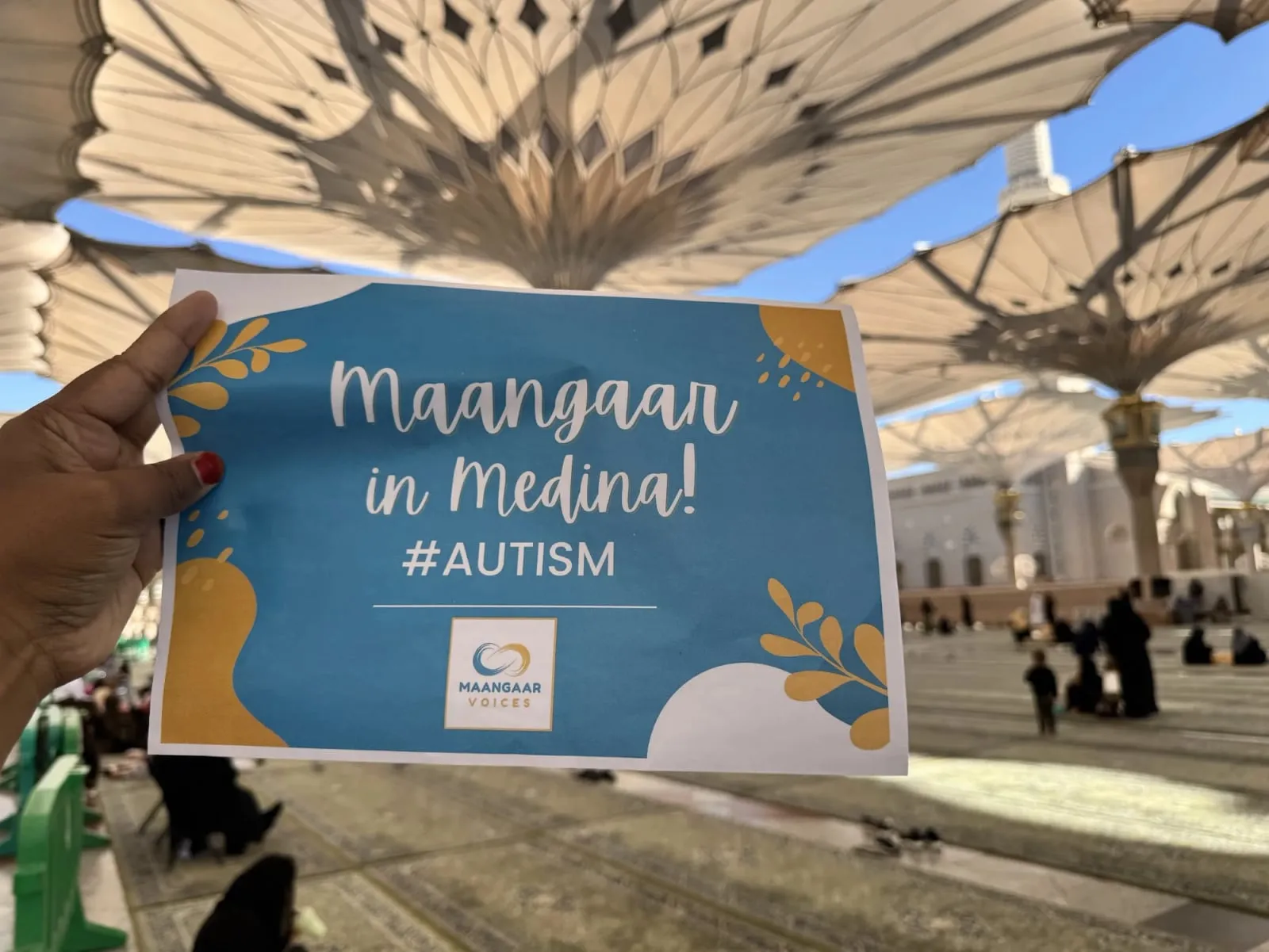 A hand holds up a blue Maangaar Voices sign reading “Maangaar in Medina! #AUTISM” beneath the white canopy umbrellas in the courtyard of Al-Masjid an-Nabawi.