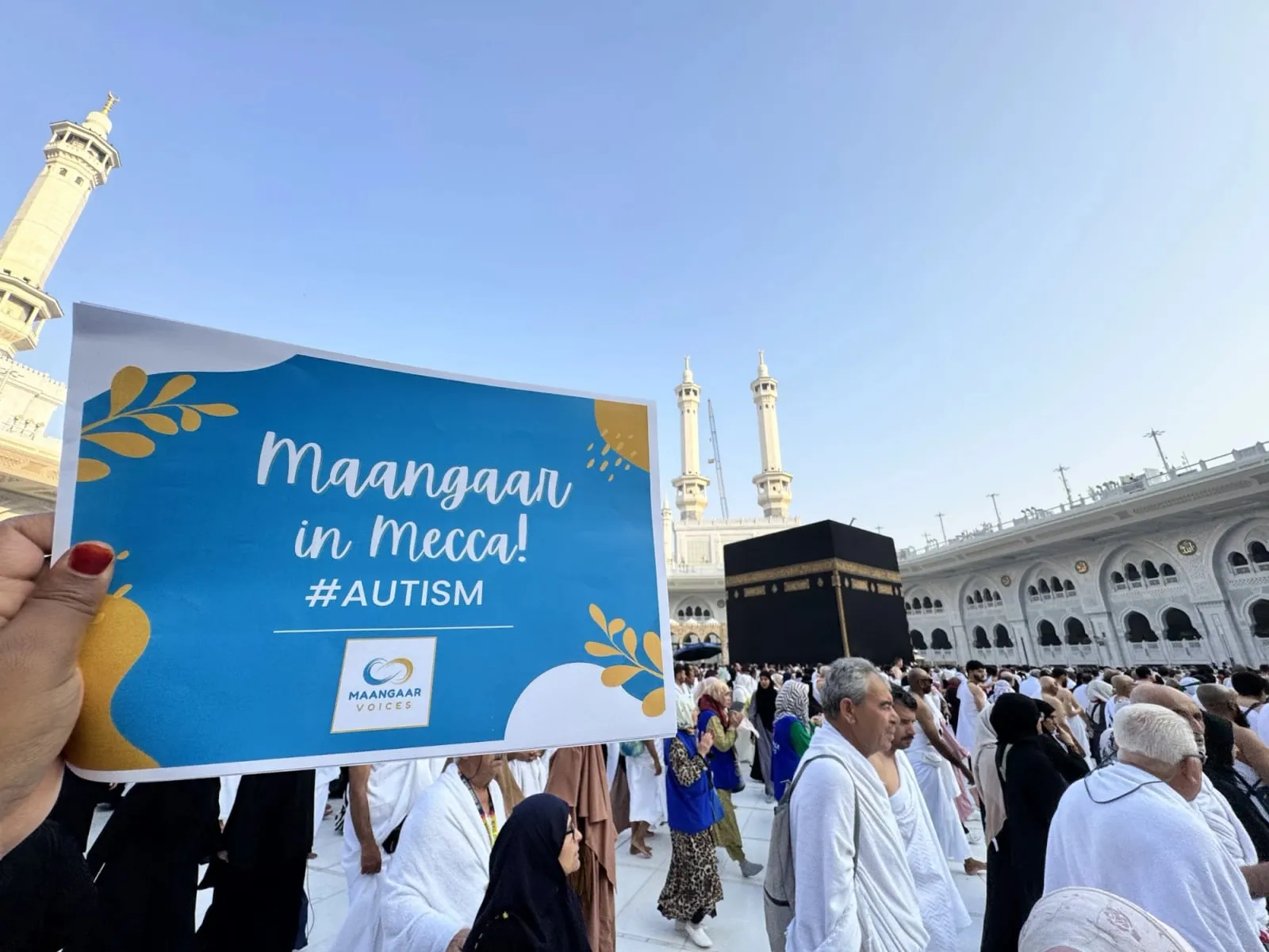 A hand holds up a blue Maangaar Voices sign reading “Maangaar in Mecca! #AUTISM” in the courtyard of Masjid al-Haram, with pilgrims in white, the minarets and the Kaaba behind it.