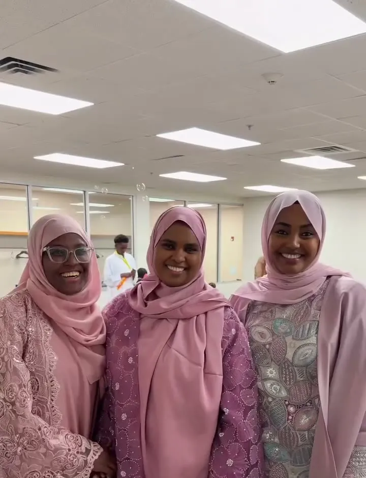 Three smiling women in pink Eid dresses inside the masjid.