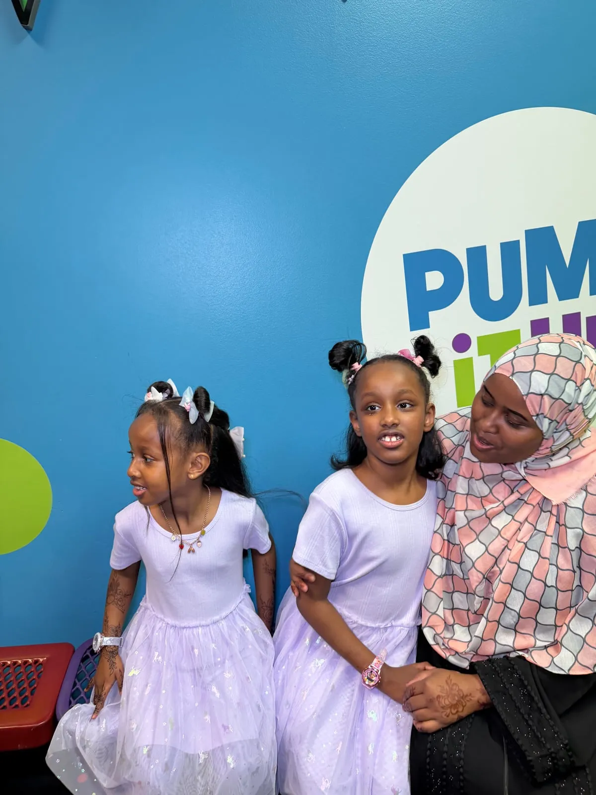 A mother in a pink-and-white checked hijab laughs with her two daughters in white Eid dresses in front of the Pump It Up sign.