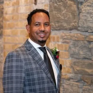 Portrait of Abdirahman Mukhtar in a checked suit and tie, in front of a stone wall.