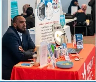 A vendor welcomes attendees at an exhibitor table.