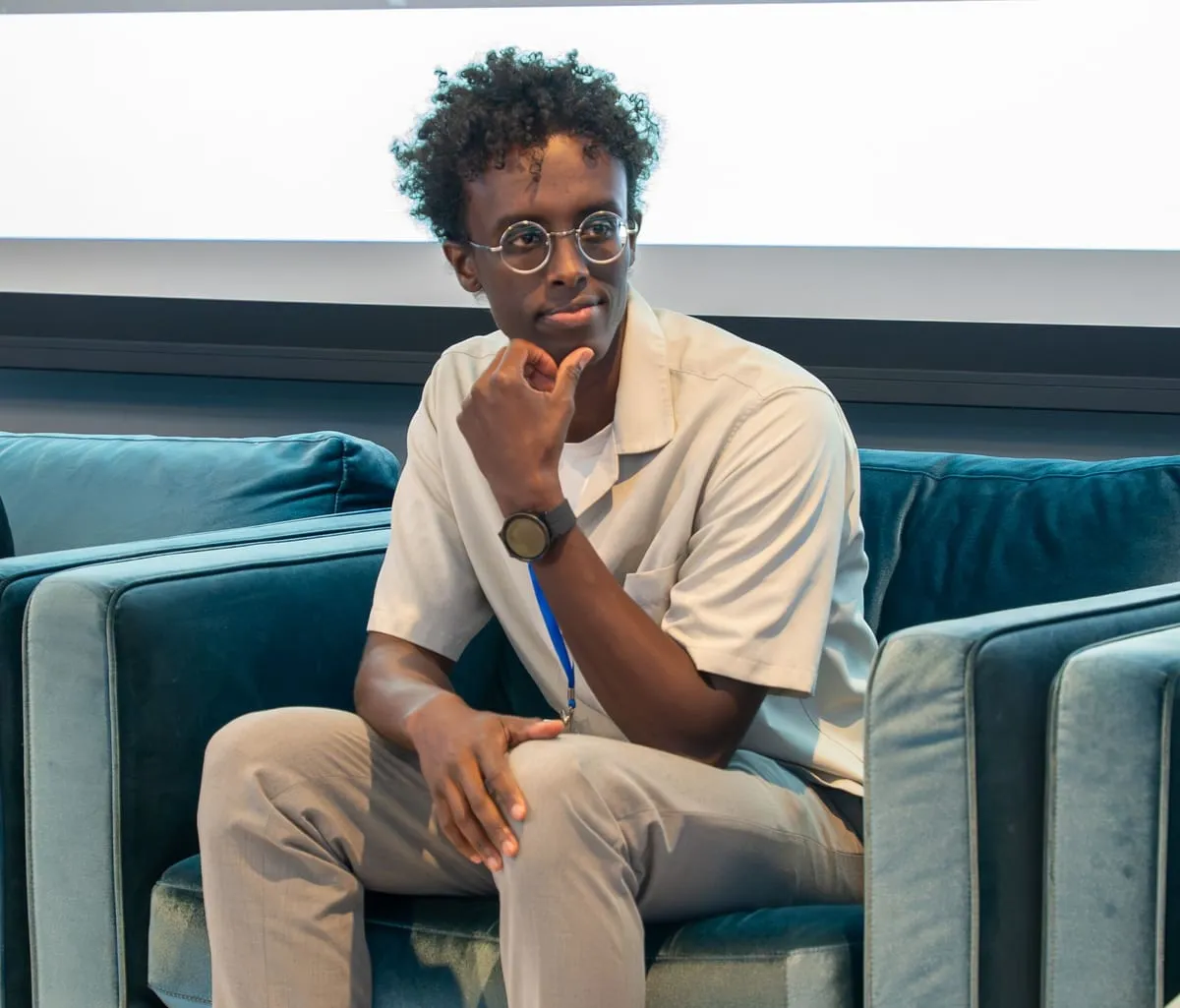A young panelist in glasses listens from a blue couch on the summit stage.