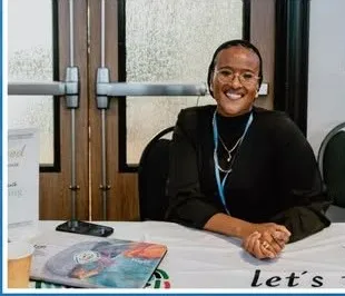 A smiling exhibitor seated at a resource table.